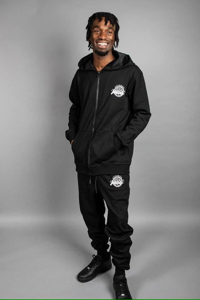 Person wearing a black hoodie and jogger set with a juice logo on chest and upper leg of jogger pant on  a gray background